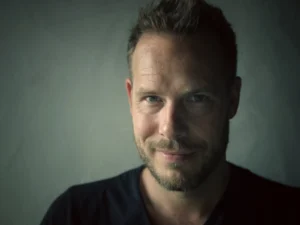 Portrait of Bas Meeuws, a man with short hair and a beard in a dark T-shirt, looking into the camera against a grey wall.