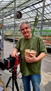 Jan C. Schlegel standing beside a large-format camera in a greenhouse, holding a potted plant.