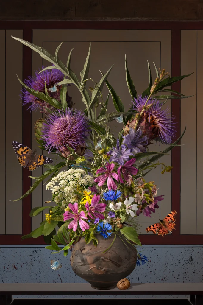 A bouquet of purple artichoke thistles, mallow, blue cornflowers and white wild carrot in a dark Roman pot on a wooden shelf, with a walnut and butterflies, against a beige Roman wall with red-brown bands.
