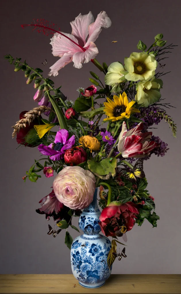 A tall bouquet of ranunculus, hibiscus and tulips in a blue-and-white Delft vase against a mauve wall.