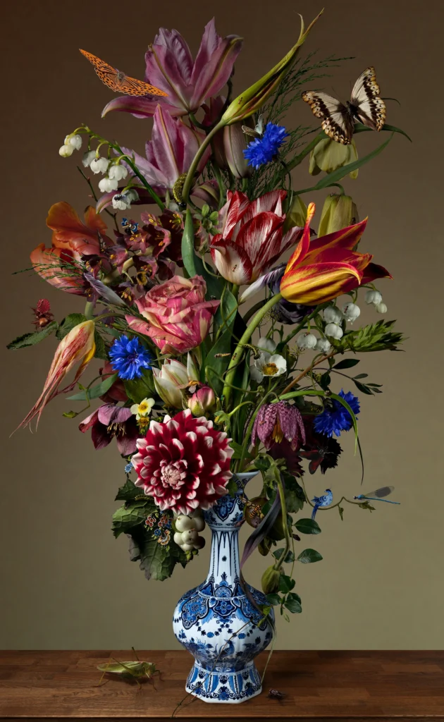 A loose bouquet of tulips, dahlias and cornflowers in a slender Delft blue vase against a grey-green wall.