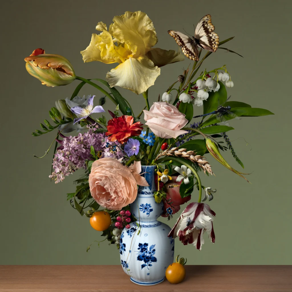 Yellow iris, tulips, a pale rose, lily of the valley and a ranunculus in a Delft blue bottle vase, with two orange tomatoes on the ledge and a swallowtail butterfly.