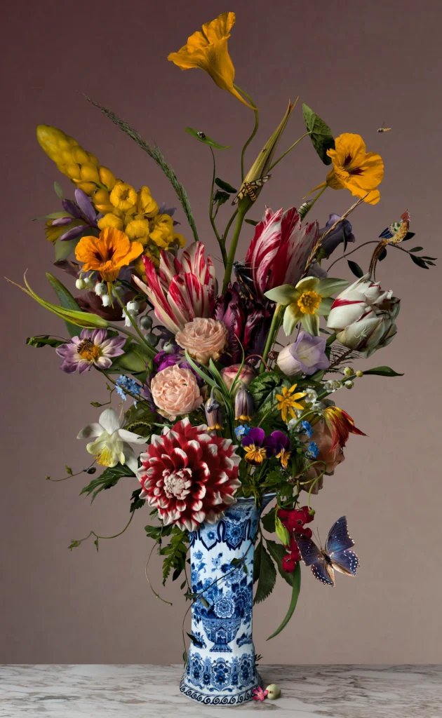 An upright bouquet of dahlias, tulips and yellow spikes in a tall Delft blue vase against a muted brown wall.