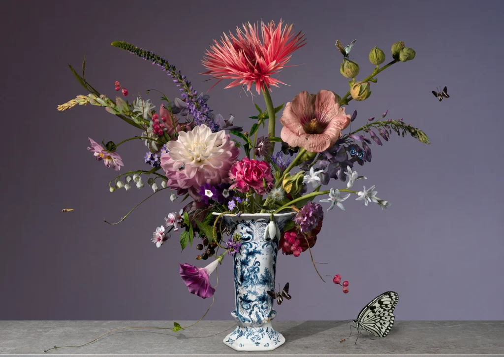A wide, airy arrangement with a fringed pink dahlia in a slim Delft blue vase on a stone ledge, with a butterfly.