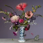 A wide, airy arrangement with a fringed pink dahlia in a slim Delft blue vase on a stone ledge, with a butterfly.