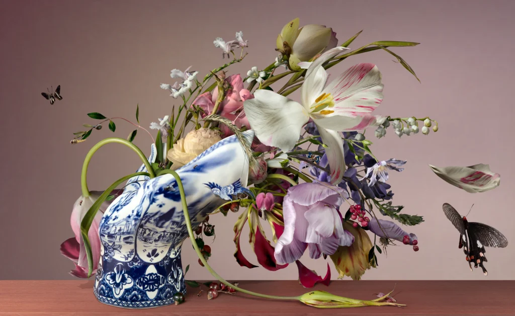 A fallen Delft blue Blow Away vase with a bouquet of tulips, lilies and peonies lying on a wooden table, with butterflies.