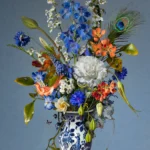 Blue delphinium, a white carnation and orange flowers with a peacock feather in a Delft blue vase, a peacock egg on the ledge.