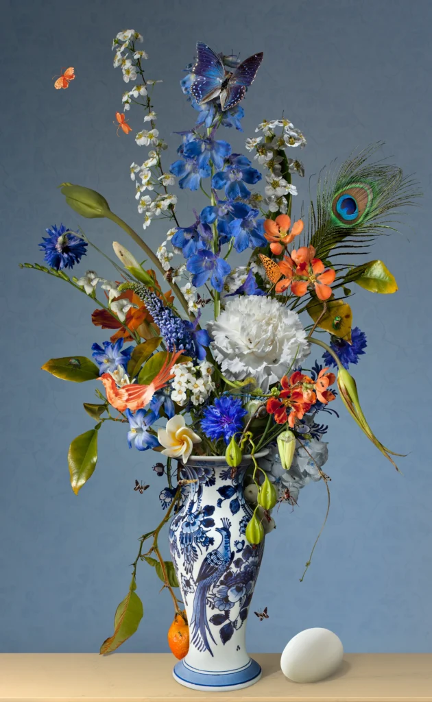 Blue delphinium, a white carnation and orange flowers with a peacock feather in a Delft blue vase, a peacock egg on the ledge.