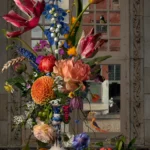 A bouquet of tulips, iris and roses in a Delft blue vase before a window, with grapes and fruit on the ledge.
