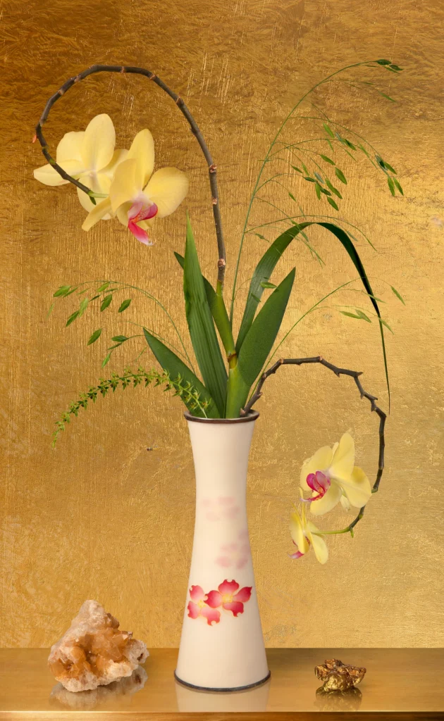 A single orchid stem with grasses in a slender white vase against a gold-leaf ground, with a piece of rose quartz on the ledge.