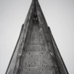 A tall triangular concrete spire narrowing to a sharp point against an empty pale sky, with a row of small openings along its edge.