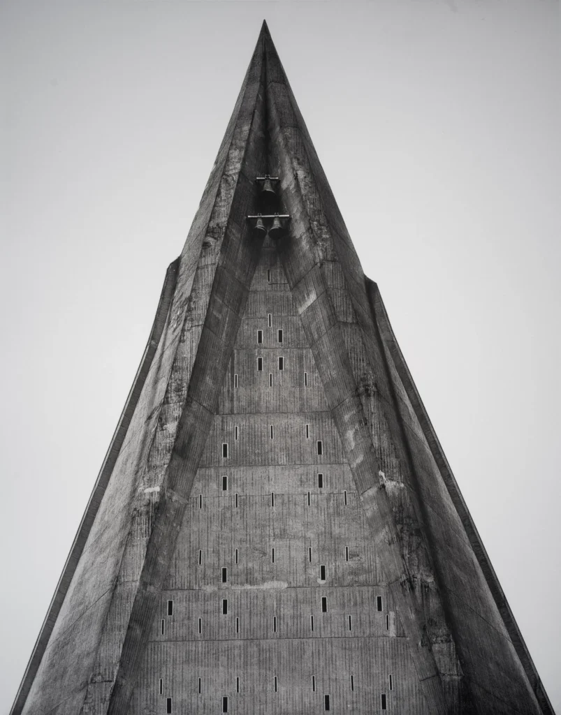 A tall triangular concrete spire narrowing to a sharp point against an empty pale sky, with a row of small openings along its edge.