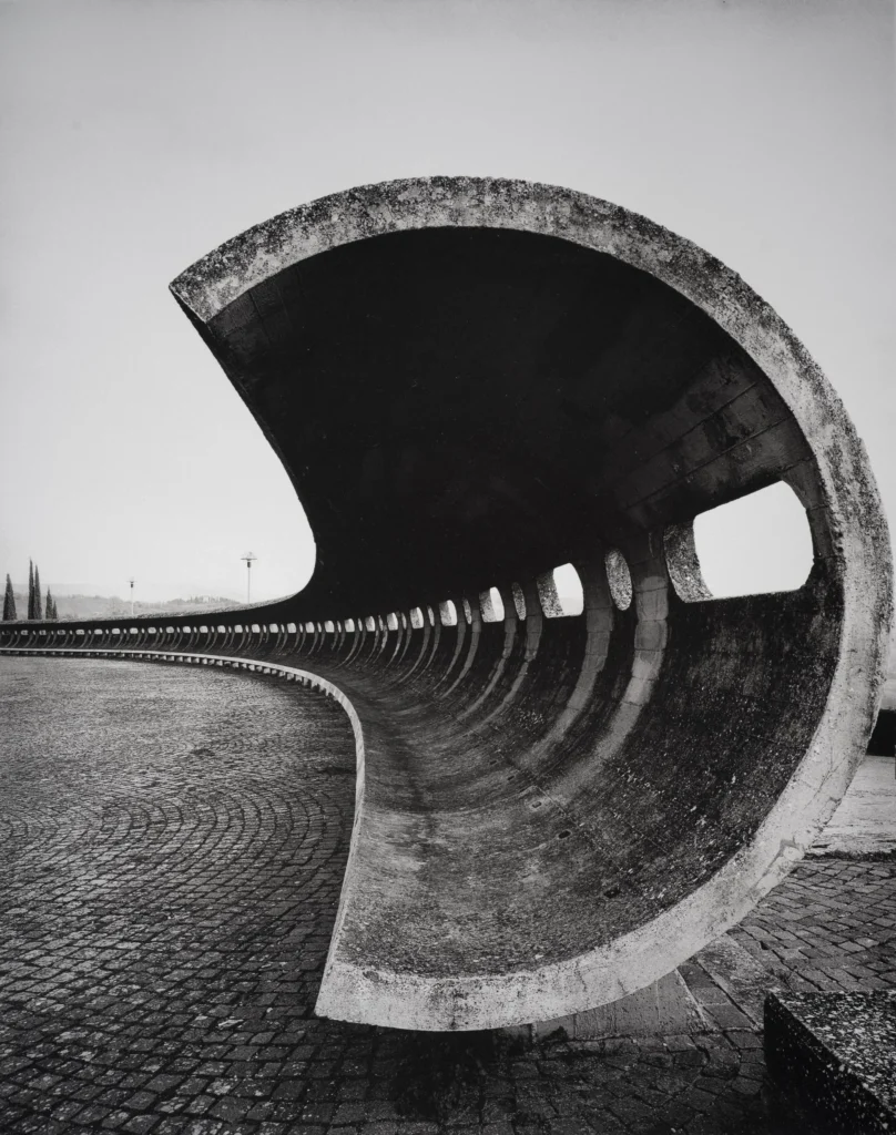 A sweeping concrete shell curving over a cobbled square, its hollow underside opening towards the viewer.