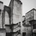 Angular concrete complex with a tall stair tower, slanted walls and overlapping blocks under a pale sky.