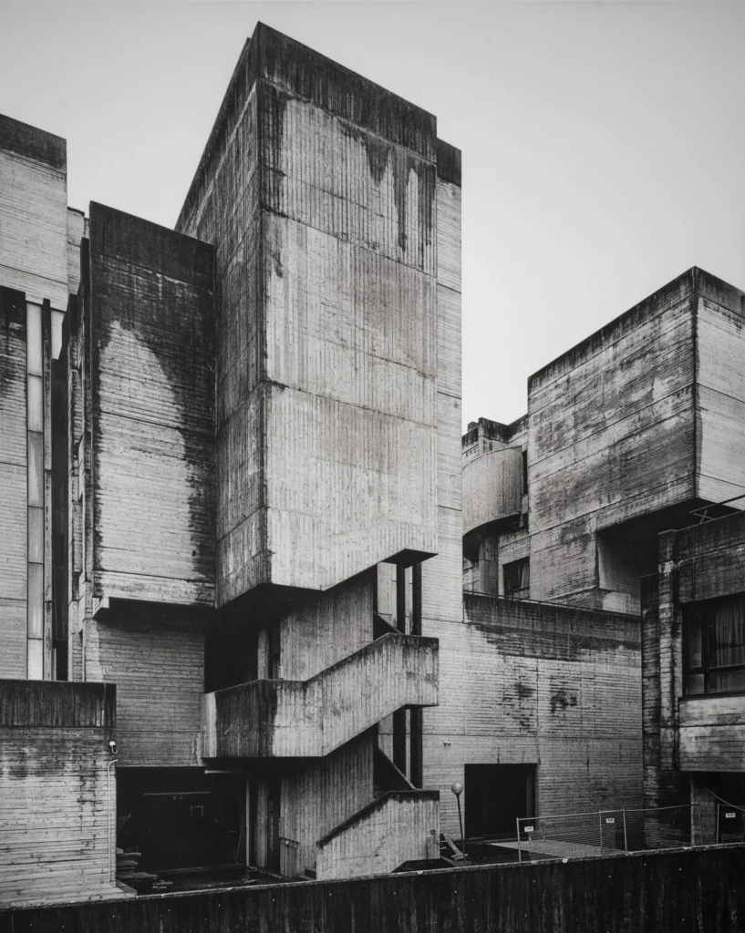 Angular concrete complex with a tall stair tower, slanted walls and overlapping blocks under a pale sky.
