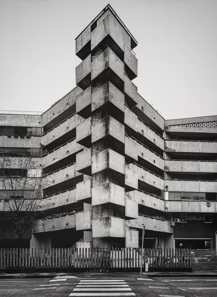 Stepped tower of stacked concrete slabs rising above a low block, with a zebra crossing in the foreground.