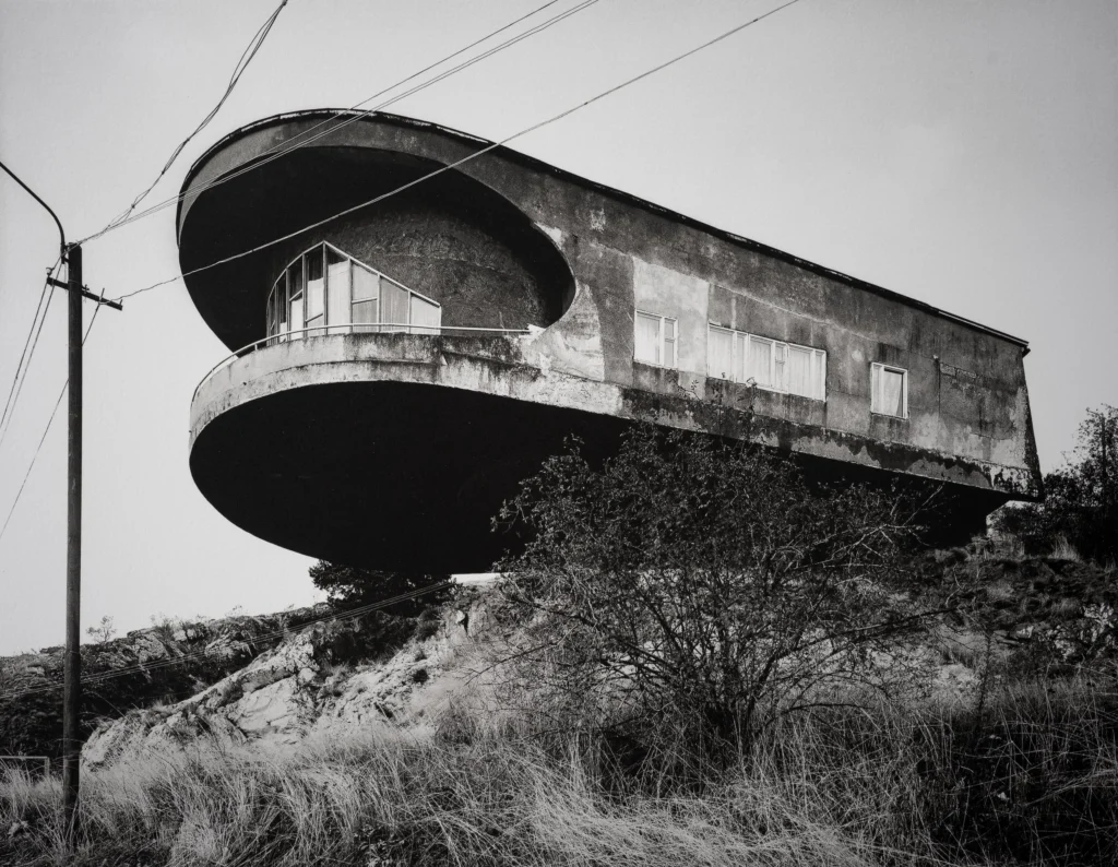 A concrete building on a hillside with an oval upper floor cantilevered far out over the slope, beside power lines.