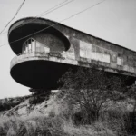 A concrete building on a hillside with an oval upper floor cantilevered far out over the slope, beside power lines.