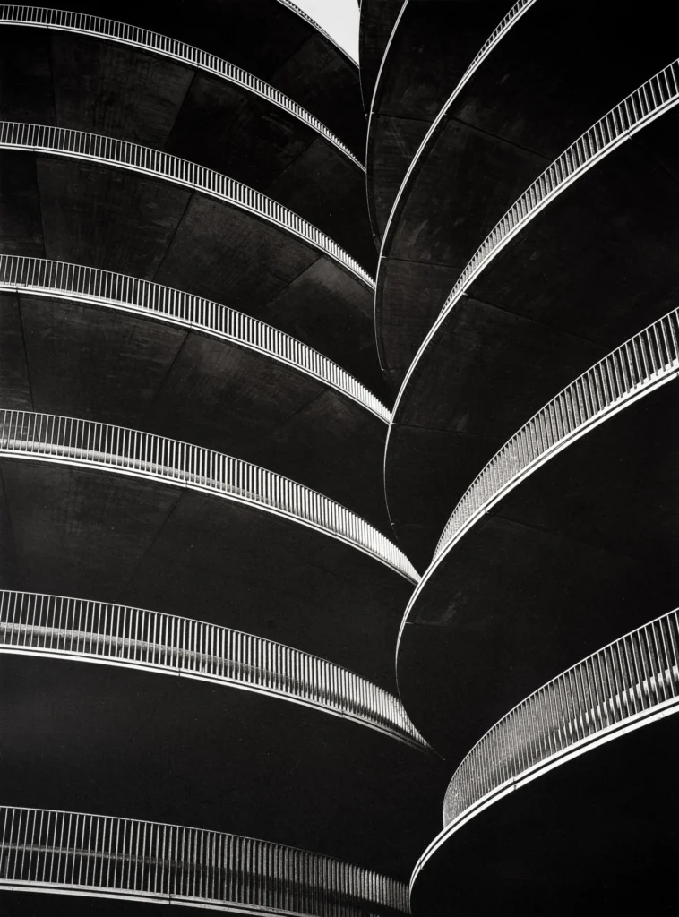 Curved concrete ramps of a spiral parking structure stacked in tight rows, their thin railings catching the light against deep shadow.
