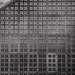 A concrete façade made of identical square windows in a dense grid, brightening towards the top and sinking into darkness below; a few pigeons sit along the sloping roof edge.