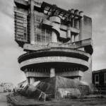 A round concrete building on a raised base with a cantilevered upper storey and a curved balcony carrying lettering, seen from below against a pale sky.
