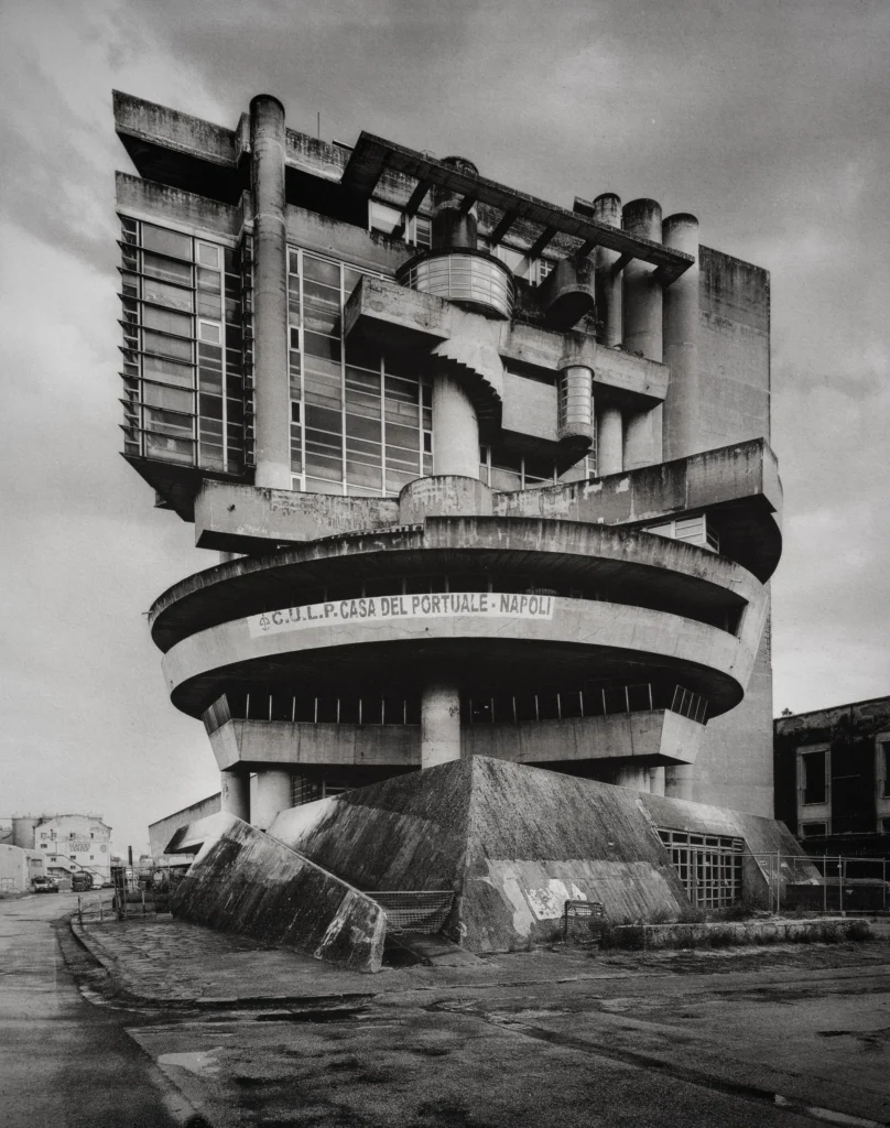 A round concrete building on a raised base with a cantilevered upper storey and a curved balcony carrying lettering, seen from below against a pale sky.