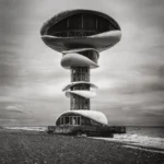 A tall concrete tower on a pebble beach carrying a saucer-shaped top storey with a band of windows and a smaller disc halfway up its stem, under a soft grey sky.