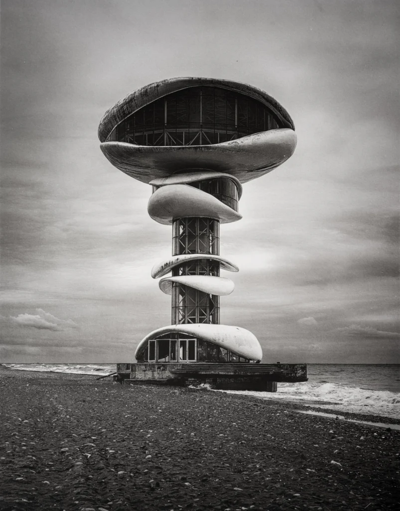 A tall concrete tower on a pebble beach carrying a saucer-shaped top storey with a band of windows and a smaller disc halfway up its stem, under a soft grey sky.
