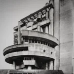 A round concrete drum with a curved wing and a stepped stair tower rising behind it, with low sheds in the foreground.