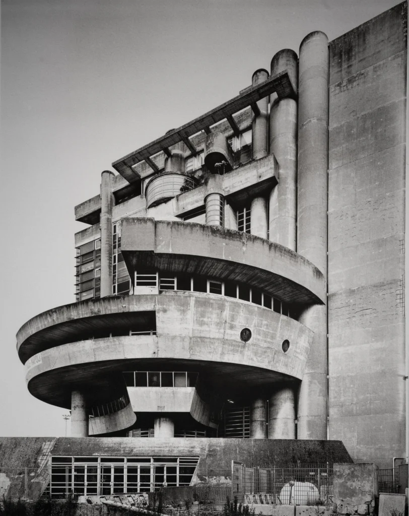 A round concrete drum with a curved wing and a stepped stair tower rising behind it, with low sheds in the foreground.