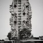 A tall slender apartment tower of irregularly stacked concrete boxes, with a bus blurred in motion at street level.