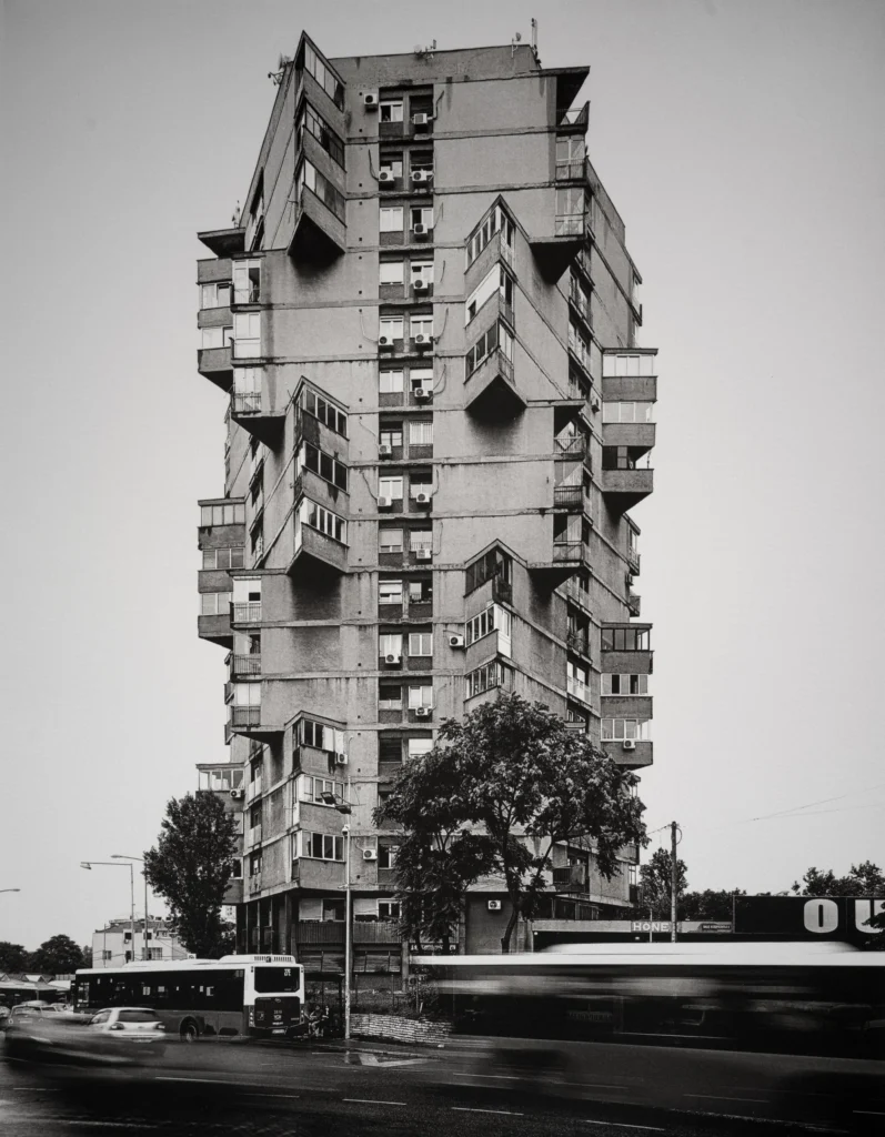 A tall slender apartment tower of irregularly stacked concrete boxes, with a bus blurred in motion at street level.