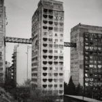 Two concrete residential towers with rows of rounded balconies, connected by a bridge structure, with a crane behind them.