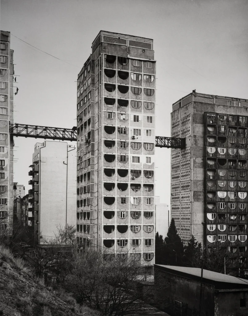 Two concrete residential towers with rows of rounded balconies, connected by a bridge structure, with a crane behind them.