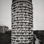 A cylindrical tower with rows of rounded openings rising from a plaza, with trees and distant blocks below.