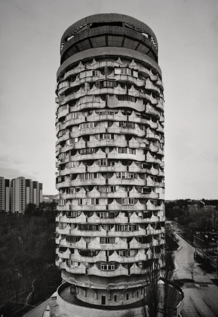 A cylindrical tower with rows of rounded openings rising from a plaza, with trees and distant blocks below.