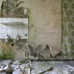 A wall with a square of white tiles, a glass shelf and two broken-off taps where a washbasin once hung; peeling green-patterned wallpaper to the right and shards of sanitary ware and plaster on the floor.